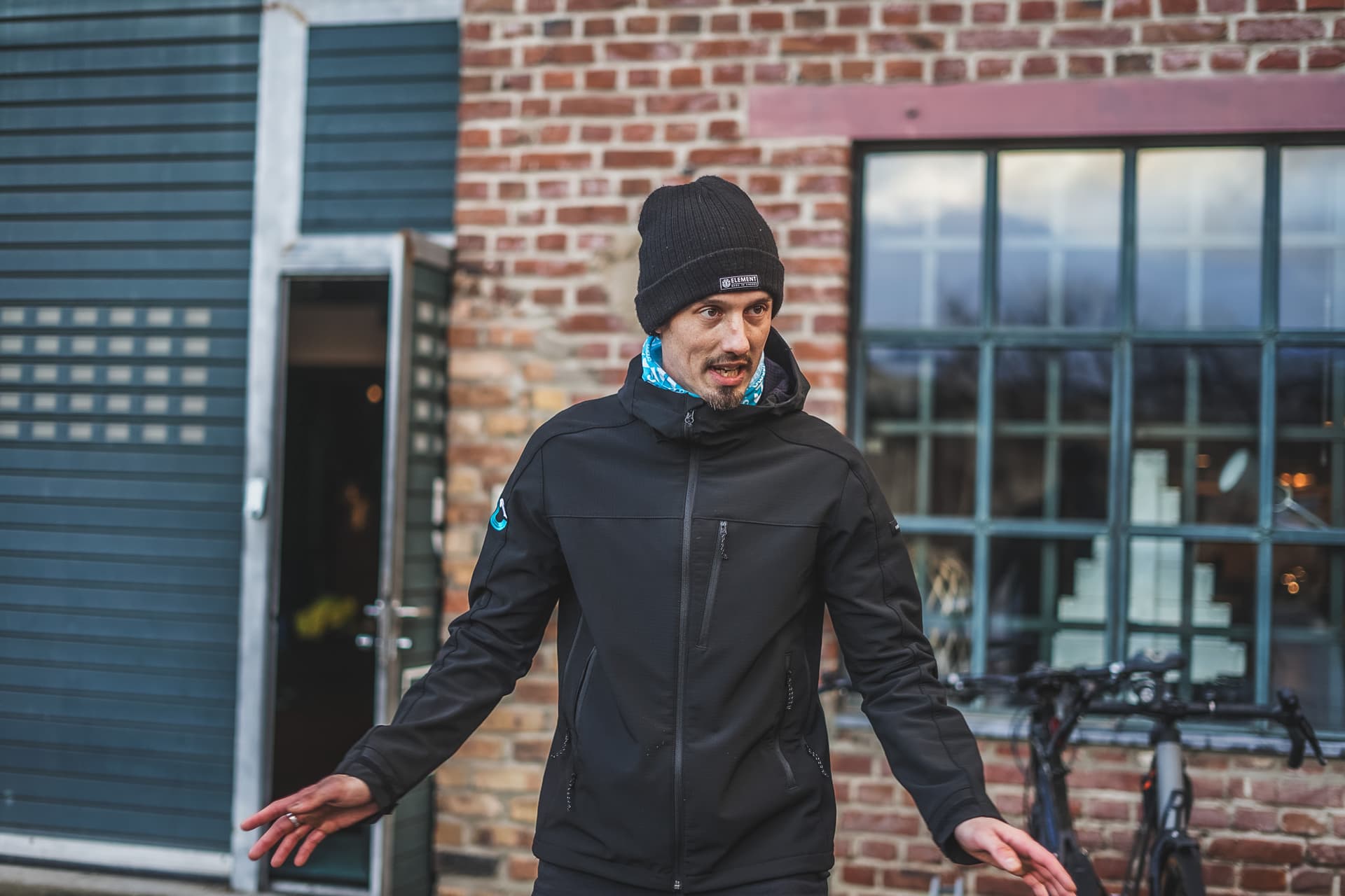 Personal Trainer Robert Biller erklärt eine Übung vor einer Backsteinwand, trägt eine schwarze Jacke und Mütze.