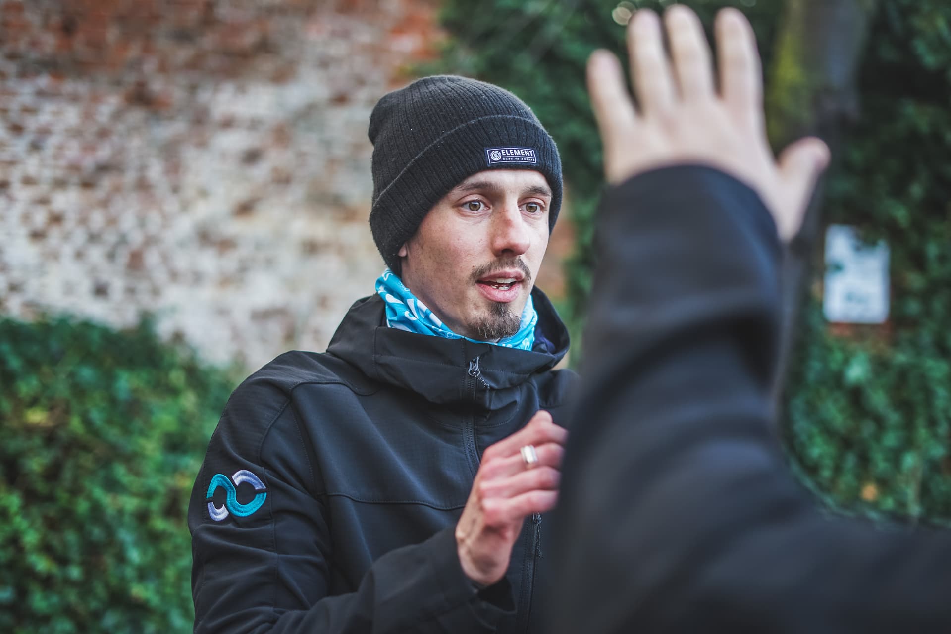 Personal Trainer Robert Biller gibt Anweisungen, mit schwarzer Mütze und Jacke, im Grünen vor einer Backsteinmauer