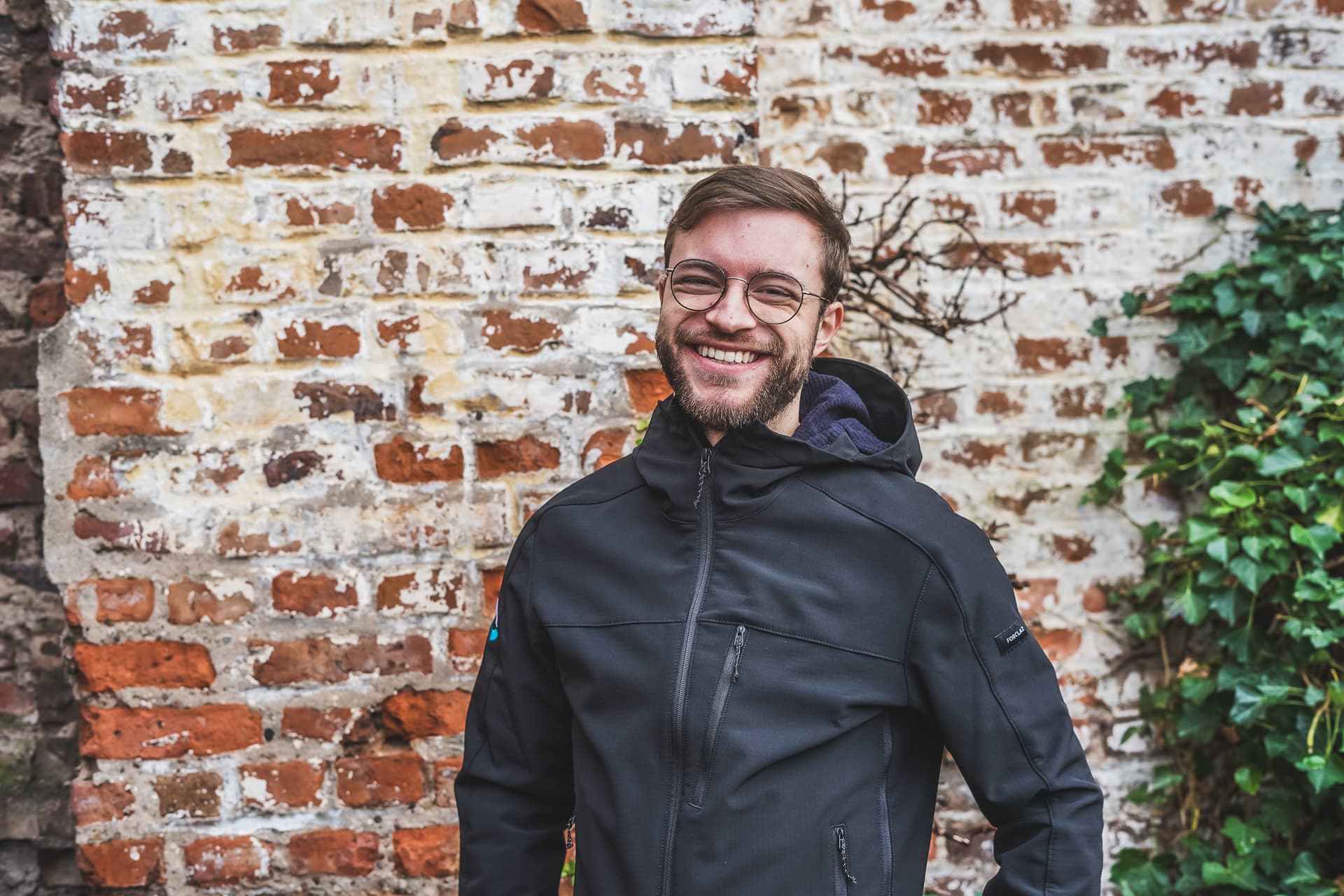 Oliver Maas steht vor einer brauen Backsteinwand und lacht herzlich in die Kamera