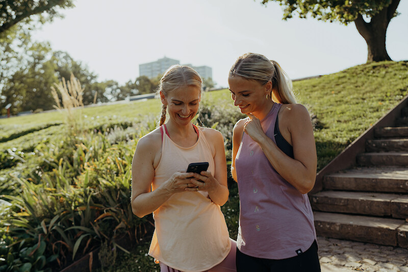 Neujahr Fitness: Zwei Frauen im Outdoor-Training schauen gemeinsam auf eine Fitness-App, Motivation durch Tracking, Fortschritt und Gemeinschaft.