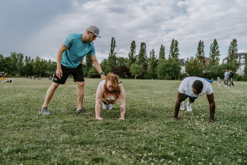 Outdoor Gym Plank-Korrektur durch Trainer – Trainer korrigiert die Haltung einer Teilnehmerin bei Plank-Übungen im Park, weiterer Athlet trainiert daneben.