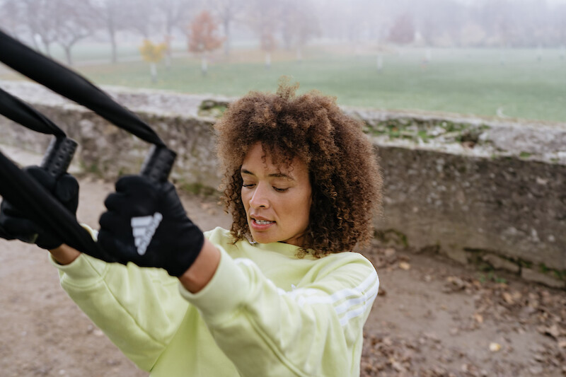 Outdoor Gym TRX-Rudern an der Mauer – Frau trainiert mit Schlingentrainer vor einer Steinmauer und führt eine Ruderbewegung im Freien aus.