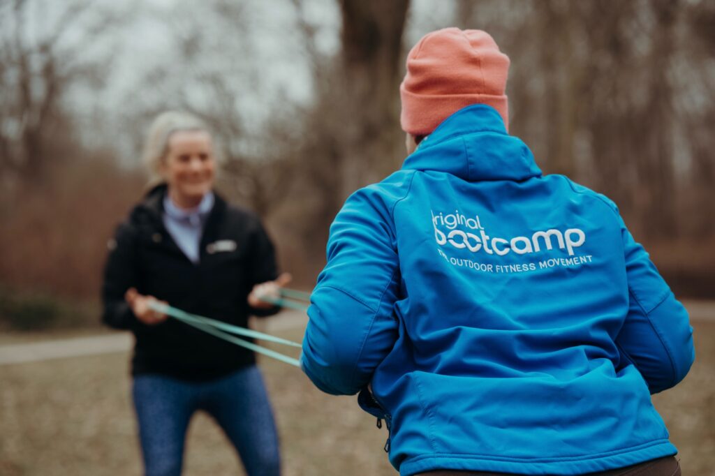 Geschenk für Sportler: Fitness-Gutschein für ein Outdoor-Bootcamp Geschenk für Sportler: Trainer in Original-Bootcamp-Jacke betreut eine Teilnehmerin bei einer Übung mit dem Widerstandsband