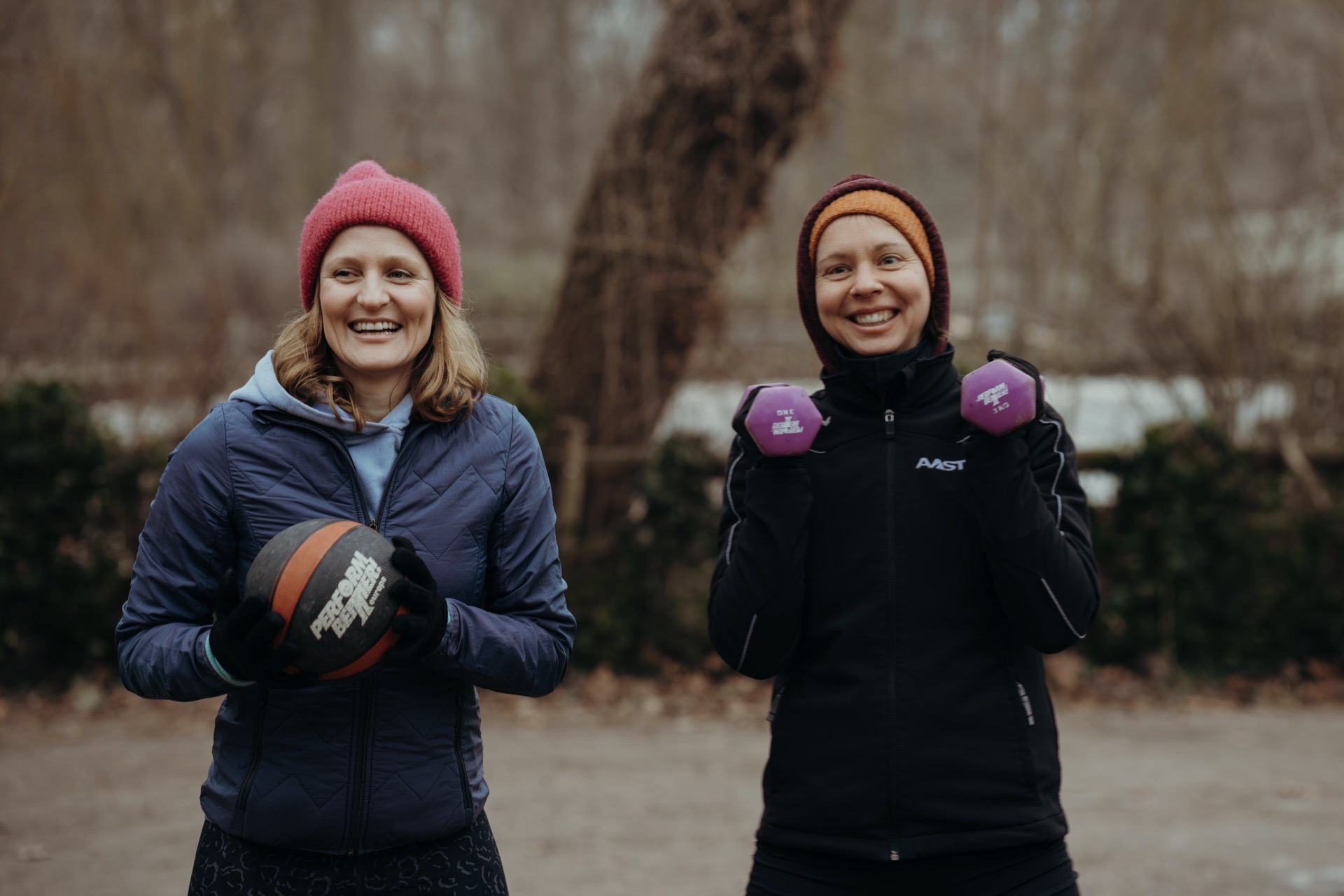 Fitness Geschenk für Frauen: Zwei lächelnde Frauen halten im winterlichen Park einen Medizinball und eine Kurzhantel