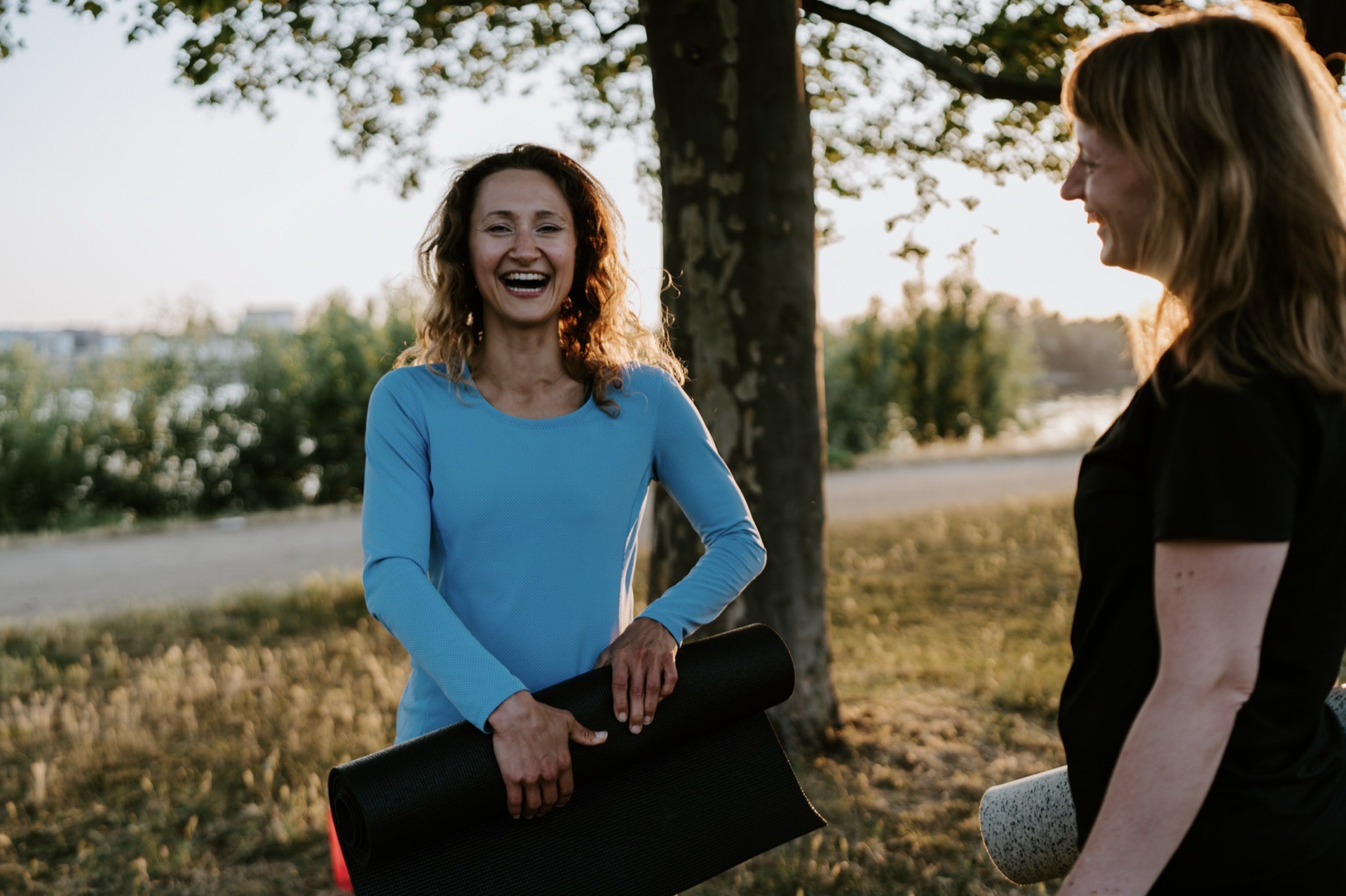Fitness Geschenk für Frauen: Eine lachende Frau hält eine Trainingsmatte unter dem Arm und spricht mit einer Freundin im Park