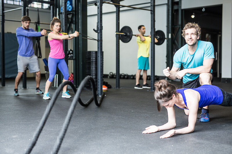 Zirkeltraining Indoor: Trainer betreut Teilnehmende bei Kraftübungen mit Battle Ropes und Langhantel.