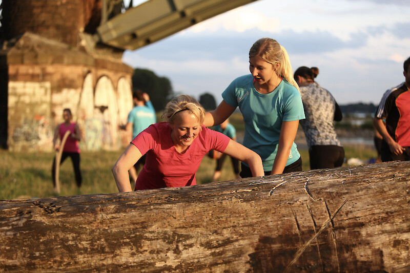 Outdoor Gym Holzstamm-Training mit Coaching – Trainerin unterstützt eine Teilnehmerin beim Überwinden eines großen Baumstamms im Freien.