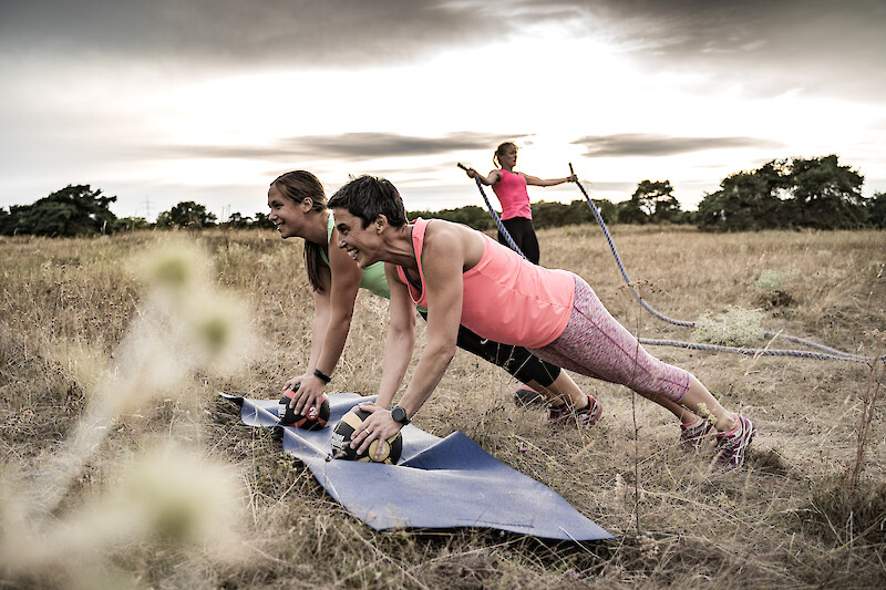 Outdoor Gym Plank mit Medizinbällen – Zwei Frauen halten eine Plank auf Medizinbällen während eine weitere Teilnehmerin im Hintergrund mit Battle Ropes trainiert.