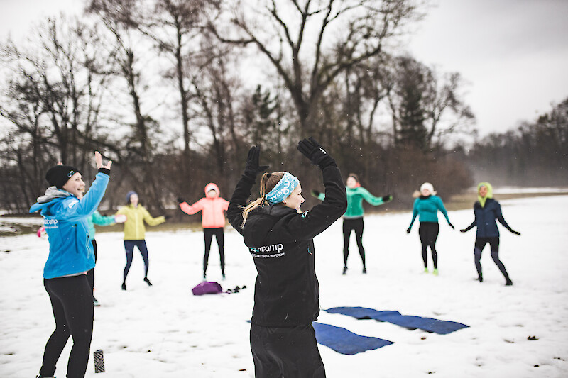 Outdoor winter training in einer motivierten Gruppe im Schnee: Trainerin leitet ein gemeinsames Outdoor-Fitness-Workout mit mehreren Teilnehmenden bei winterlichen Temperaturen im Park.