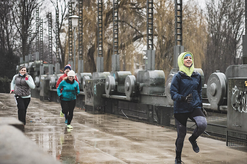 Outdoor winter training in der Stadt: Lauf- und Intervalltraining einer Gruppe bei Regen und Kälte auf einer Brücke, angeleitet von einer Trainerin im Winter-Outfit