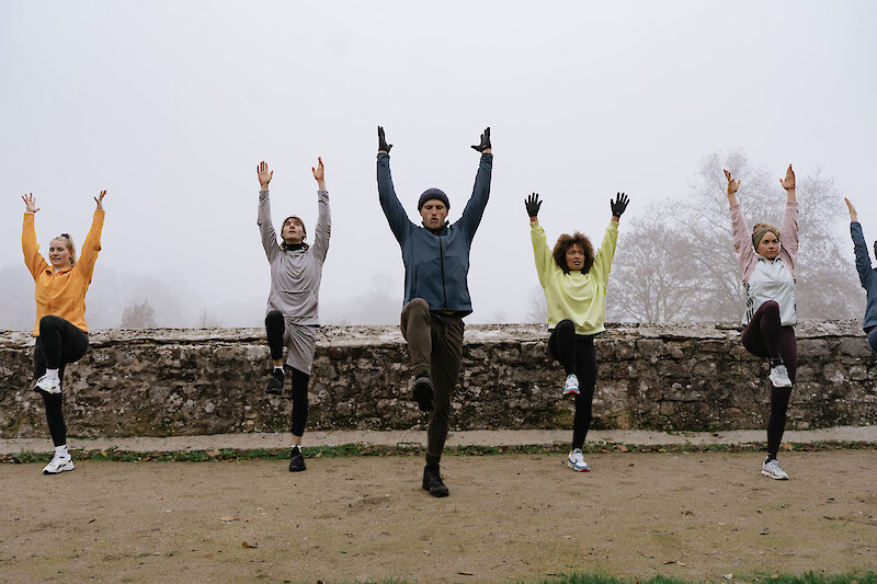 Outdoor Fitness Kiel: Gruppe beim funktionellen Training im Freien, Gleichgewichtsübung mit erhobenem Bein und Armen in einem Park bei kühlem, nebligem Wetter.