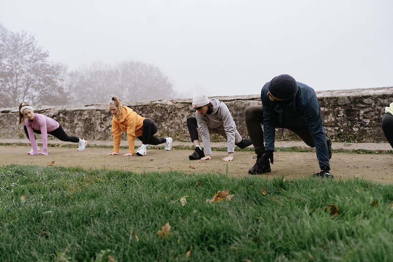 Outdoor Fitness Kiel: Gruppe beim funktionellen Outdoor-Training im Park, Ausfallschritt-Übung auf Grasfläche bei kühlem, nebligem Wetter.